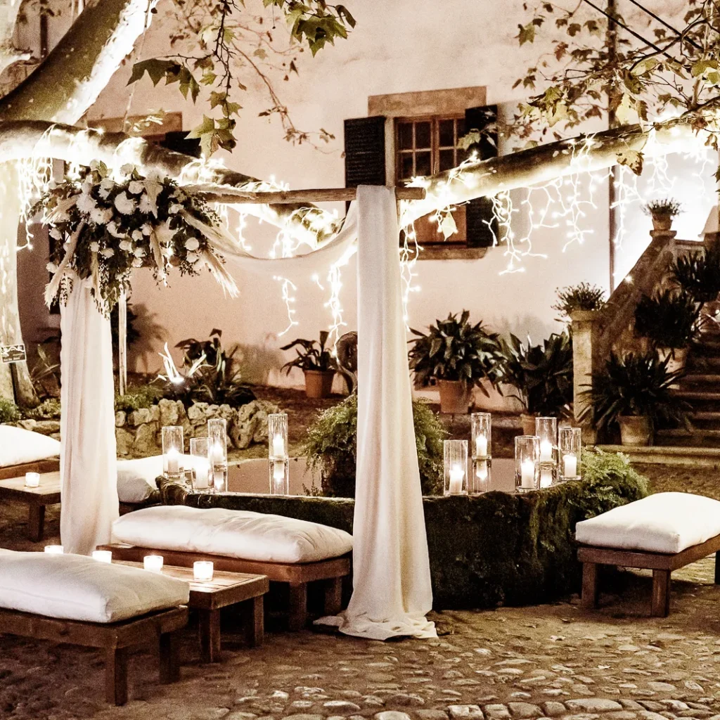Outdoor wedding ceremony setup under a large tree with white cushion seating, candles, and floral décor