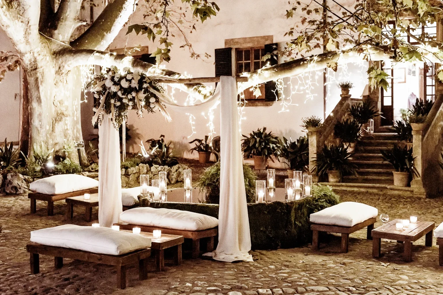Outdoor wedding ceremony setup under a large tree with white cushion seating, candles, and floral décor